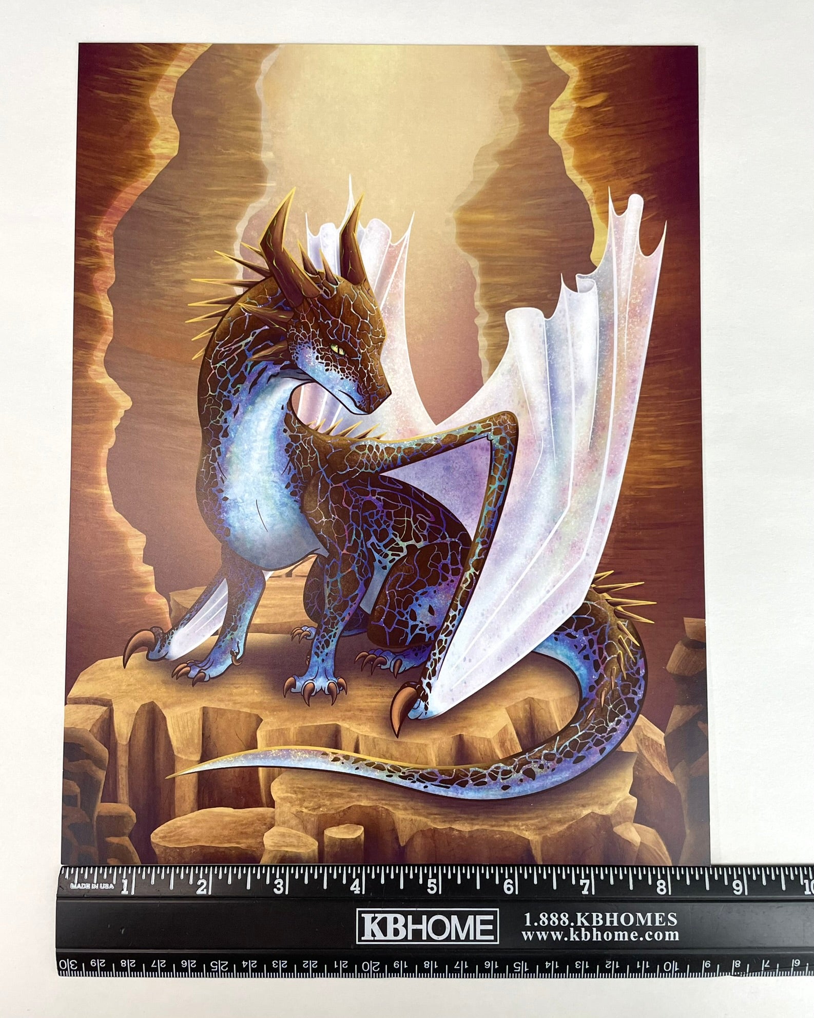 A 8 1/2 x 11 art print featuring a sitting, side profile of a dragon that has the features of a boulder opal with hues of blue, green, purple, and pink on the underbelly and cracks of the rough, brown hue, rock scales. The wings are tucked back and are of an iridescent shimmery hue. The background of the print is that of a mesa canyon with the dragon sitting on a large boulder within the canyon.