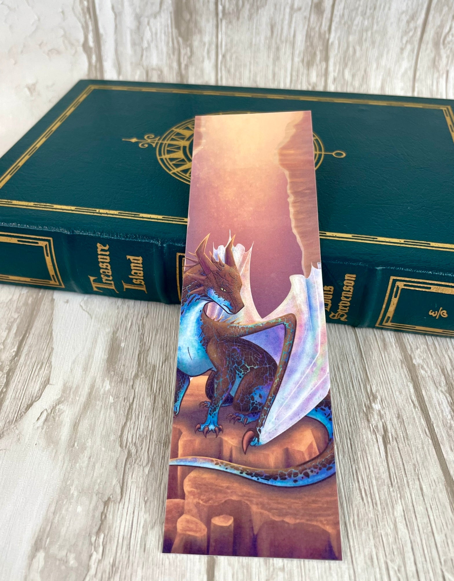 A 2 by 7 inch bookmark featuring a sitting, side profile of a dragon that has the features of a boulder opal with hues of blue, green, purple, and pink on the underbelly and cracks of the rough, brown hue, rock scales. The wings are tucked back and are of an iridescent shimmery hue that glitters in the light.  The background of the print is that of a mesa canyon with the dragon sitting on a large boulder within the canyon.