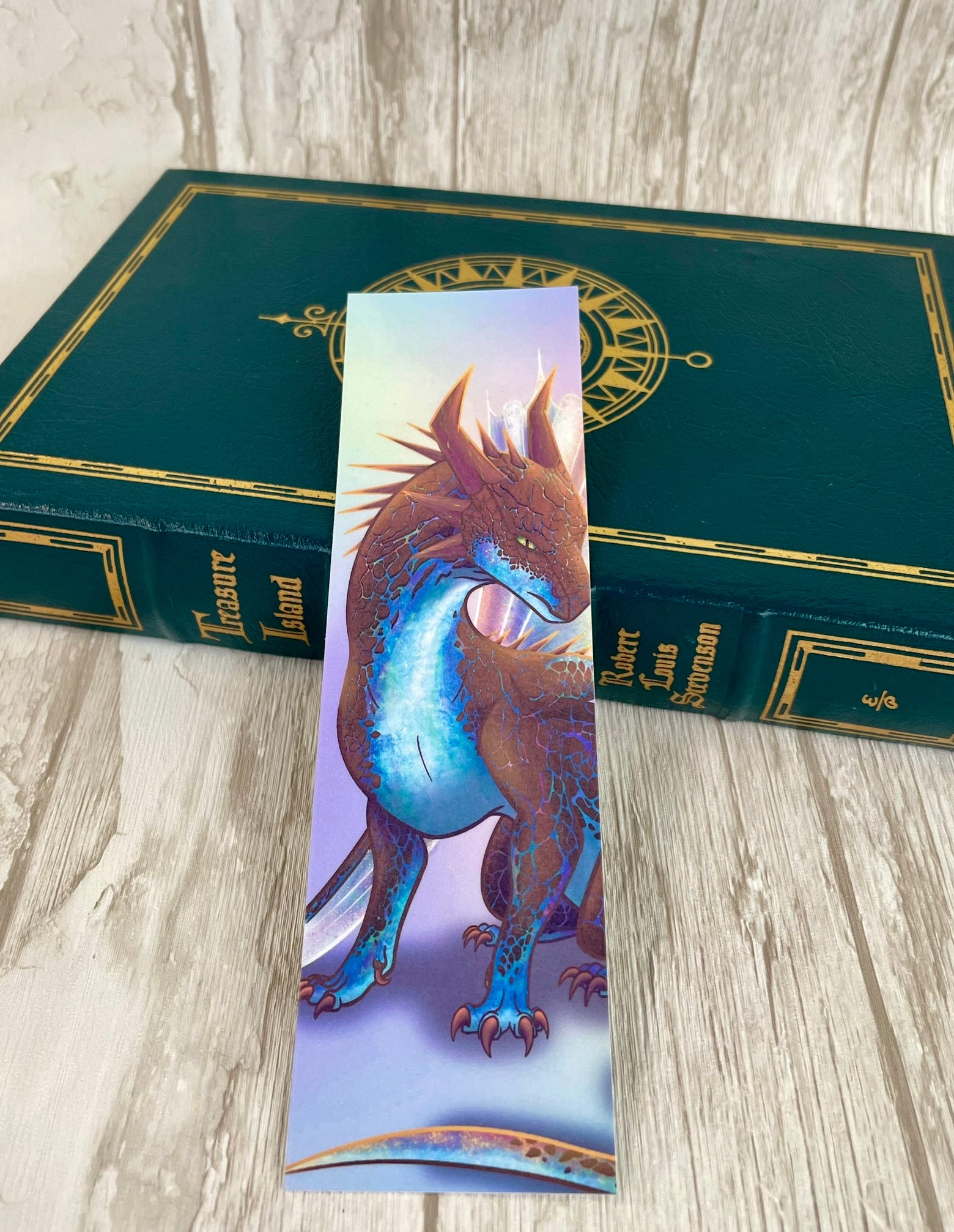 A 2 by 7 inch bookmark featuring a sitting, side profile of a dragon that has the features of a boulder opal with hues of blue, green, purple, and pink on the underbelly and cracks of the rough, brown hue, rock scales. The wings are tucked back and are of an iridescent shimmery hue that glitters in the light.  The background is of a rainbow ombre effect