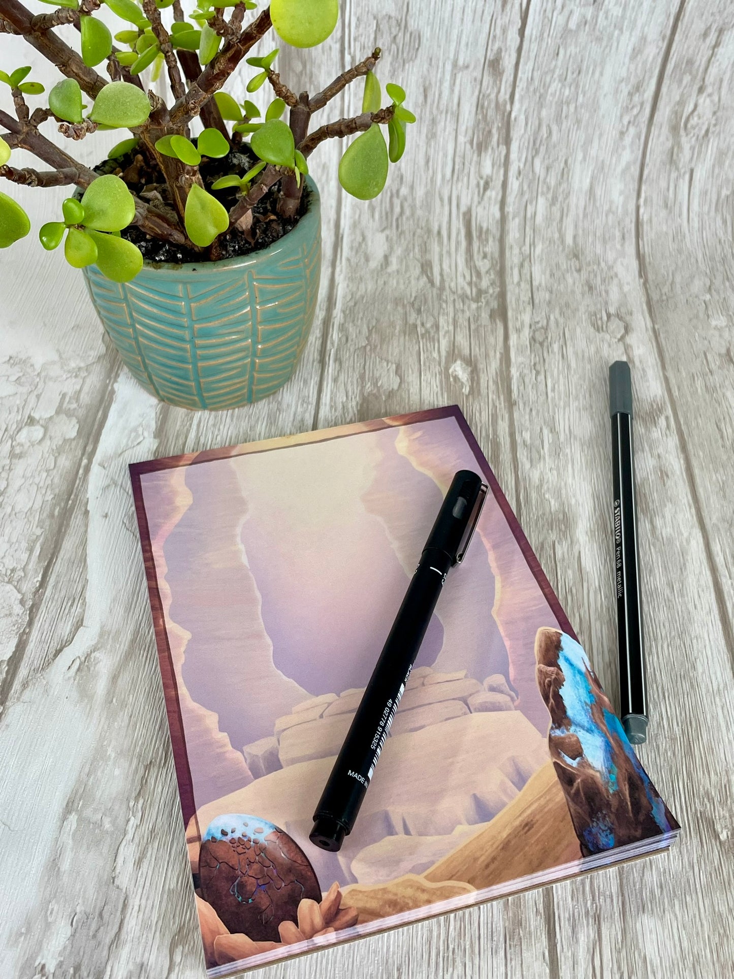 A 5 by 7 inch notepad with 50 pages that each feature the mesa canyon that is the background from the art print. Each page background is blank and shows off the opal dragon egg in the left bottom hand corner and a large raw opal mineral rock on the bottom right hand corner.