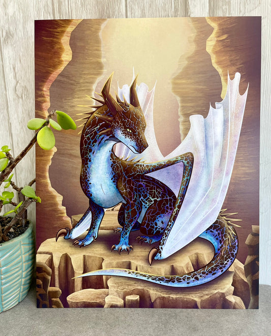 A 8 1/2 x 11 art print featuring a sitting, side profile of a dragon that has the features of a boulder opal with hues of blue, green, purple, and pink on the underbelly and cracks of the rough, brown hue, rock scales. The wings are tucked back and are of an iridescent shimmery hue.  The background of the print is that of a mesa canyon with the dragon sitting on a large boulder within the canyon.