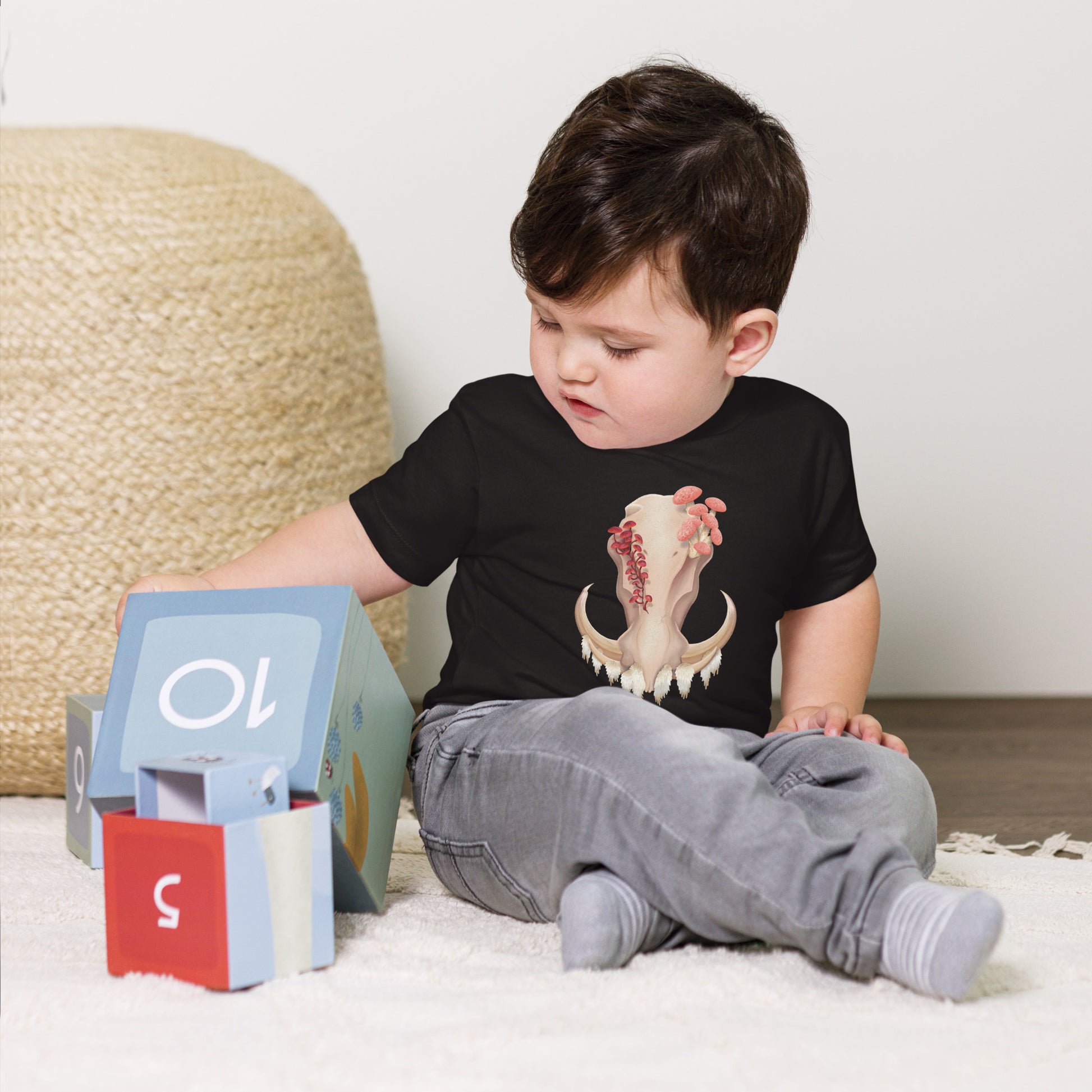 A baby sitting on the floor playing with toys wearing a black T shirt featuring a front facing warthog skull with 3 different kinds of fungi growing out of it