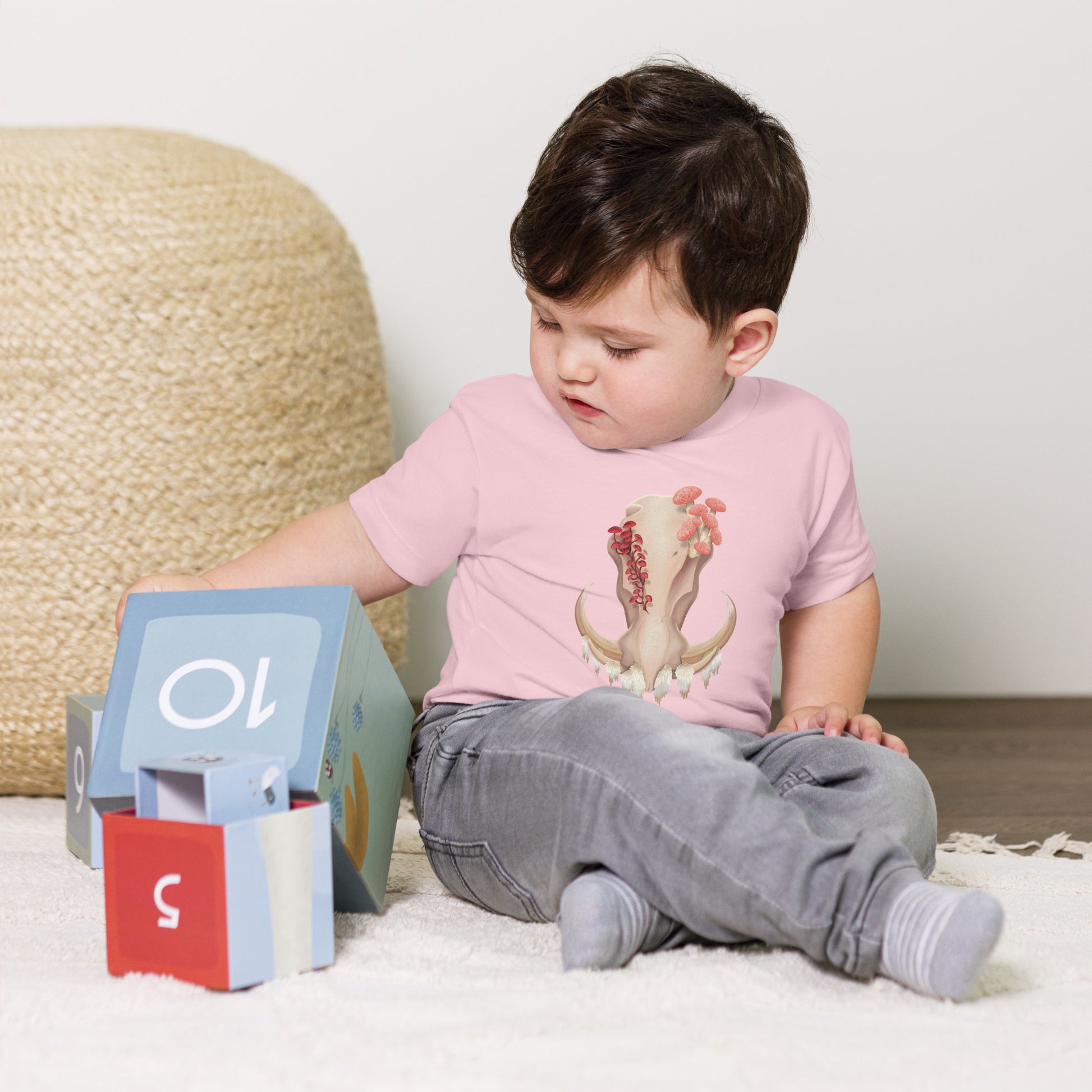 A baby sitting on the floor playing with toys wearing a light pink T shirt featuring a front facing warthog skull with 3 different kinds of fungi growing out of it