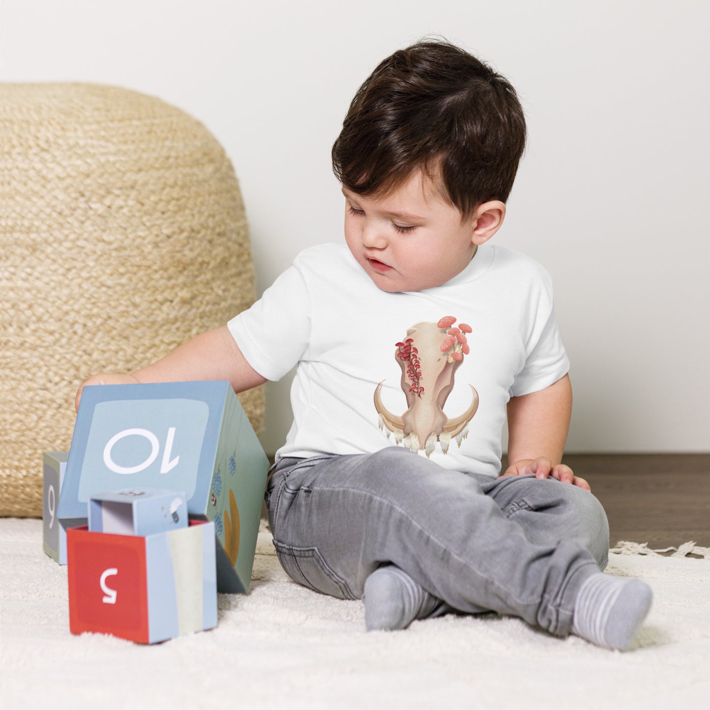 A baby sitting on the floor playing with toys wearing a white T shirt featuring a front facing warthog skull with 3 different kinds of fungi growing out of it