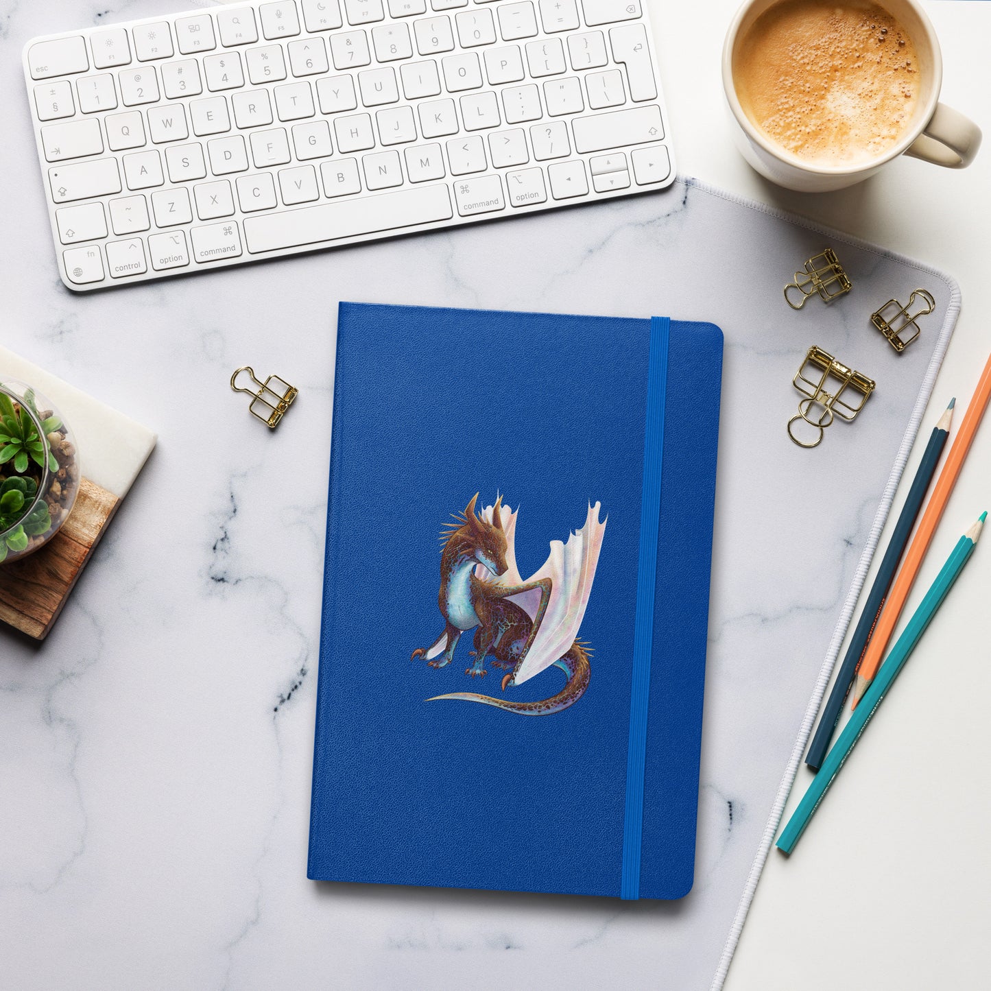 A royal blue notebook with an elastic closure, 80 lined pages, a ribbon bookmark, and pocket in the back with the front featuring a sitting, side profile of a dragon that has the features of a boulder opal with hues of blue, green, purple, and pink on the underbelly and cracks of the rough, brown hue, rock scales. The wings are tucked back and are of an iridescent shimmery hue