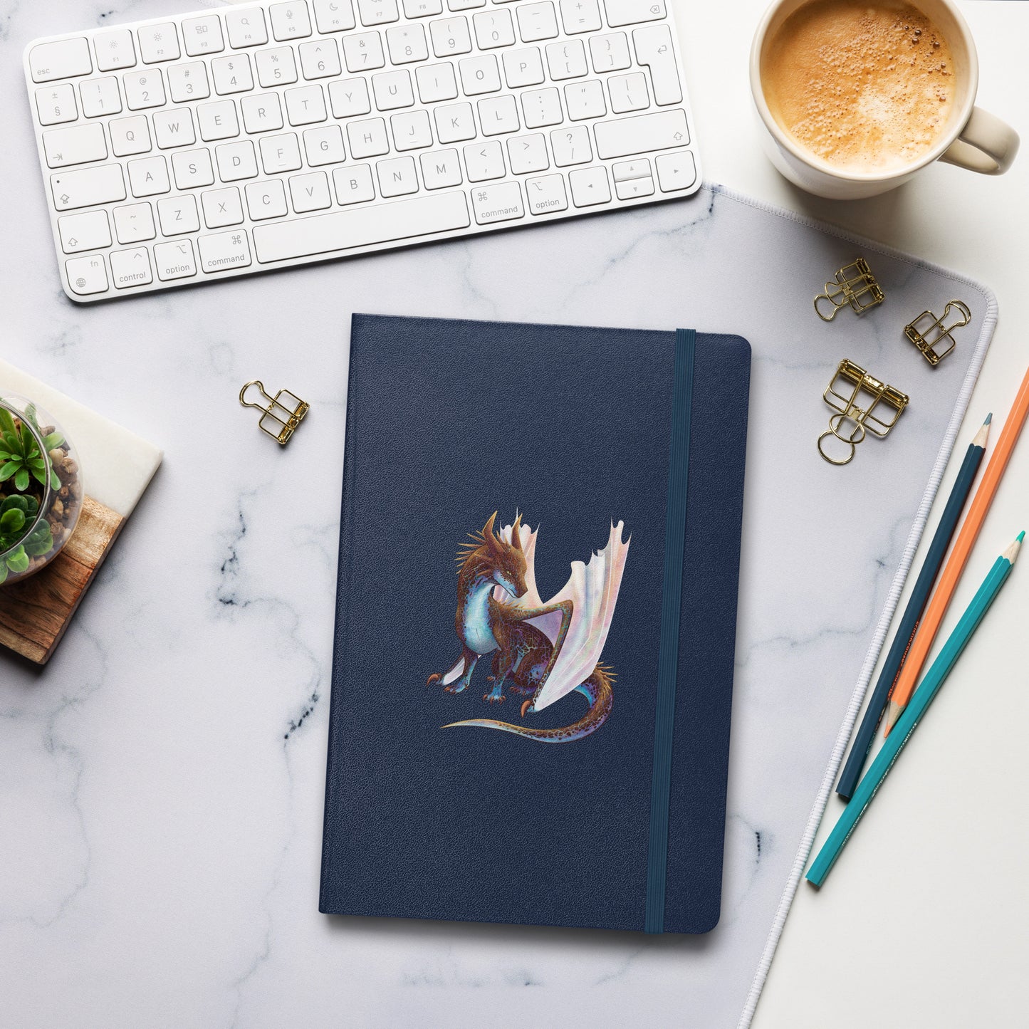 A navy blue notebook with an elastic closure, 80 lined pages, a ribbon bookmark, and pocket in the back with the front featuring a sitting, side profile of a dragon that has the features of a boulder opal with hues of blue, green, purple, and pink on the underbelly and cracks of the rough, brown hue, rock scales. The wings are tucked back and are of an iridescent shimmery hue