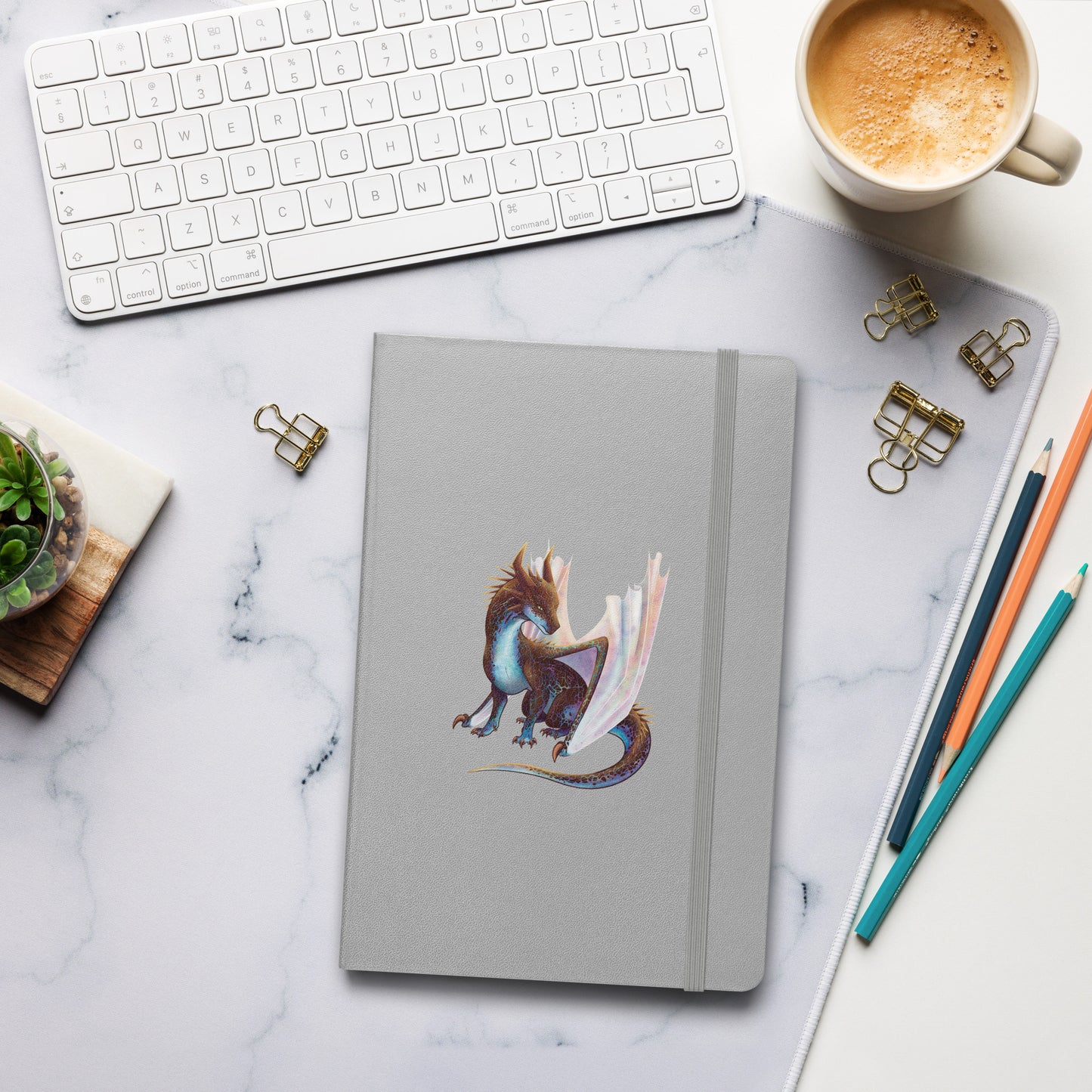 A silver notebook with an elastic closure, 80 lined pages, a ribbon bookmark, and pocket in the back with the front featuring a sitting, side profile of a dragon that has the features of a boulder opal with hues of blue, green, purple, and pink on the underbelly and cracks of the rough, brown hue, rock scales. The wings are tucked back and are of an iridescent shimmery hue