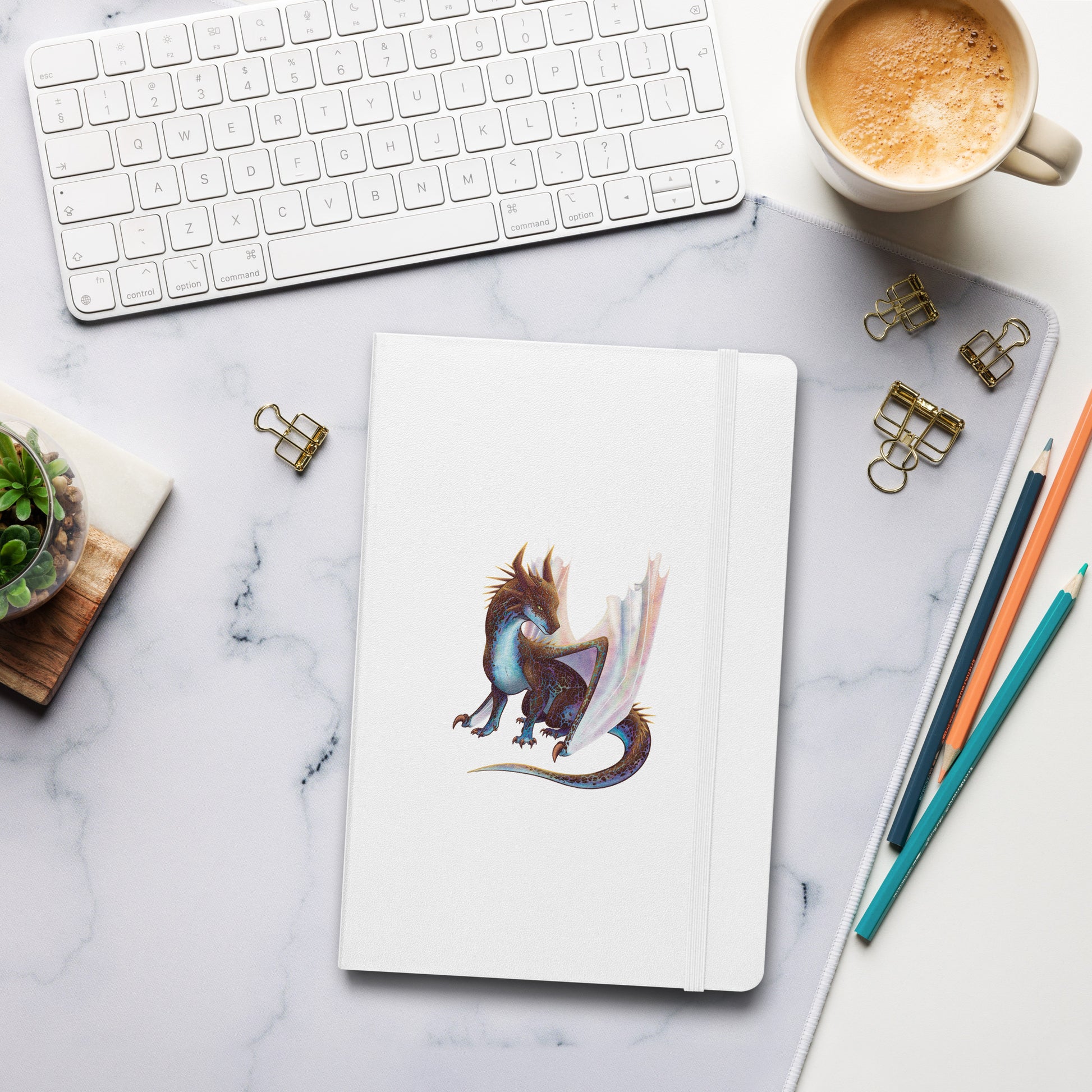 A white notebook with an elastic closure, 80 lined pages, a ribbon bookmark, and pocket in the back with the front featuring a sitting, side profile of a dragon that has the features of a boulder opal with hues of blue, green, purple, and pink on the underbelly and cracks of the rough, brown hue, rock scales. The wings are tucked back and are of an iridescent shimmery hue