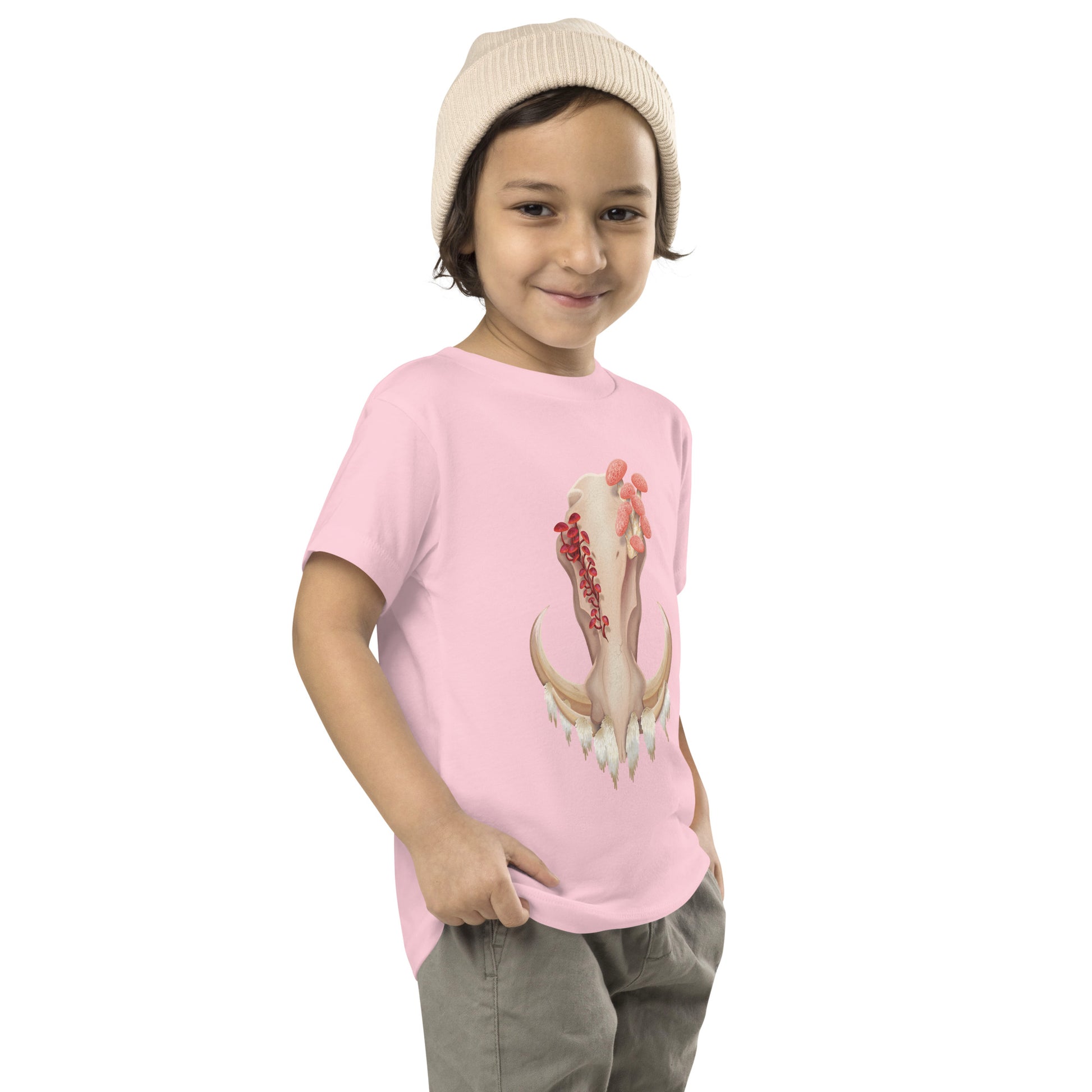 A toddler wearing a light pink T shirt featuring a front facing warthog skull with 3 different kinds of fungi growing out of it